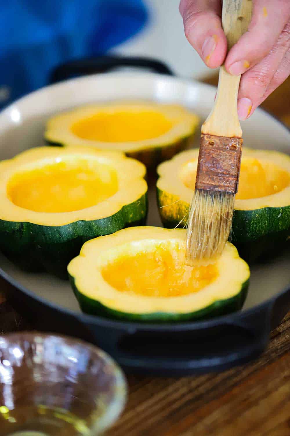 A hand brushing olive oil onto acorn squash that has been cut in half and sitting in a baking dish.