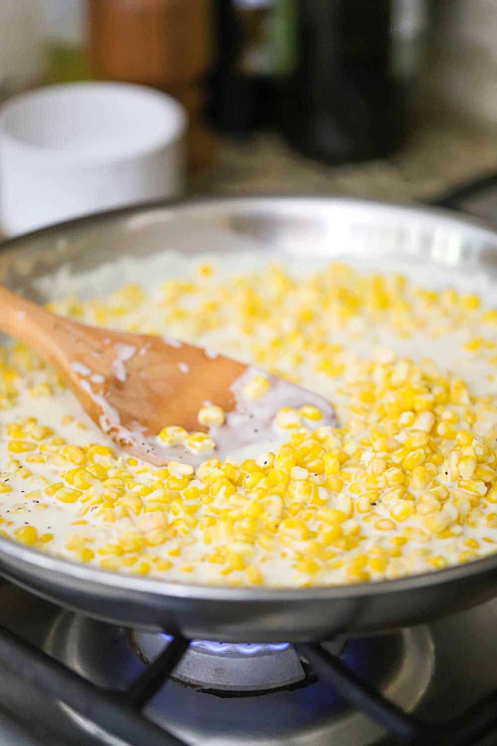 A large silver skillet on the stove filled with homemade creamed corn with a wooden spoon in it.