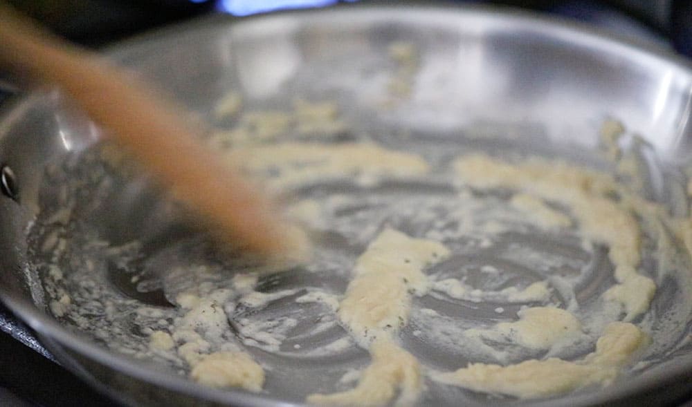 A stainless steel skillet filled with a blonde roux with a wooden spoon stirring it.