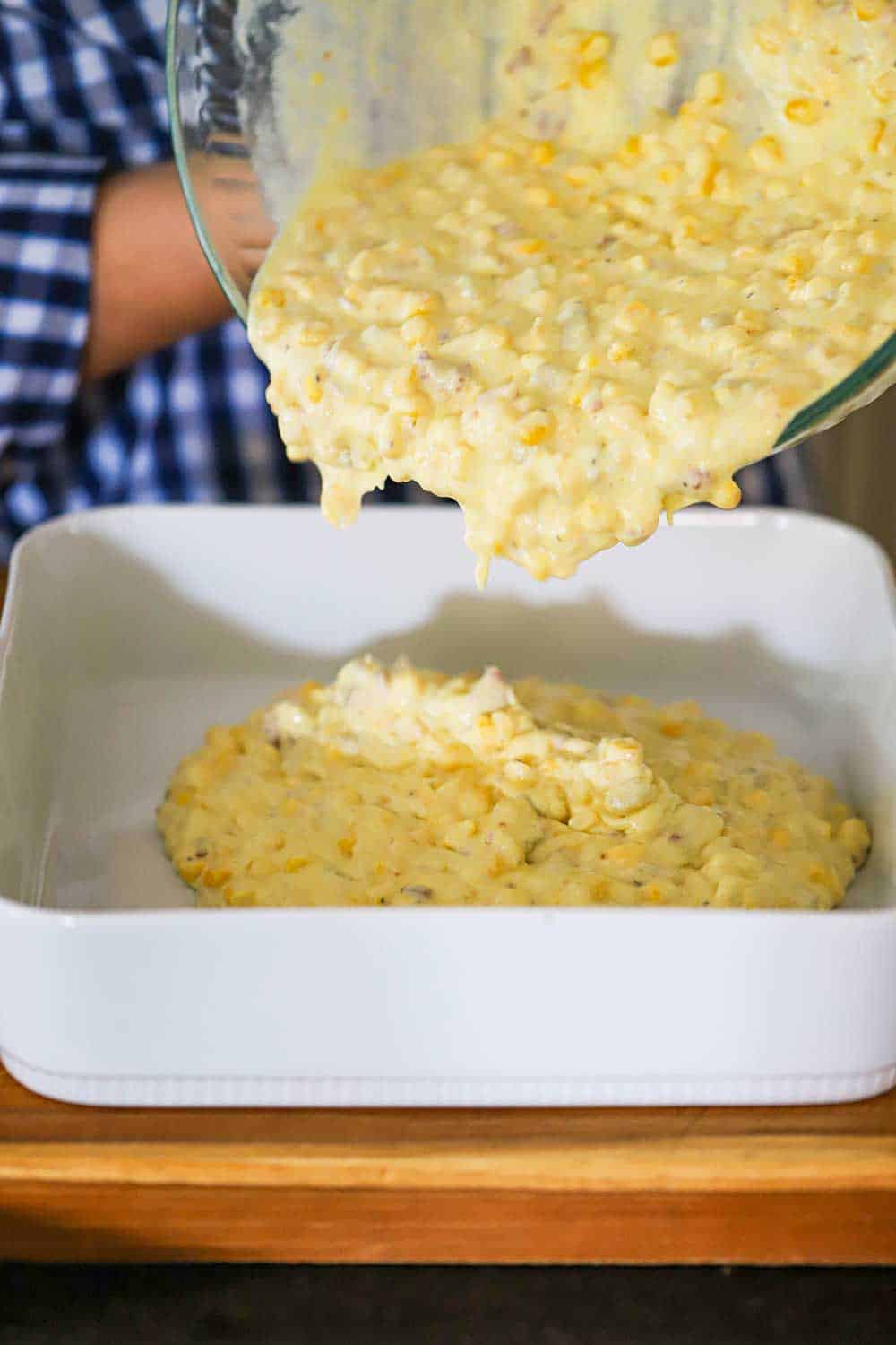 A person pouring a corn casserole uncooked mixture into a square baking dish.
