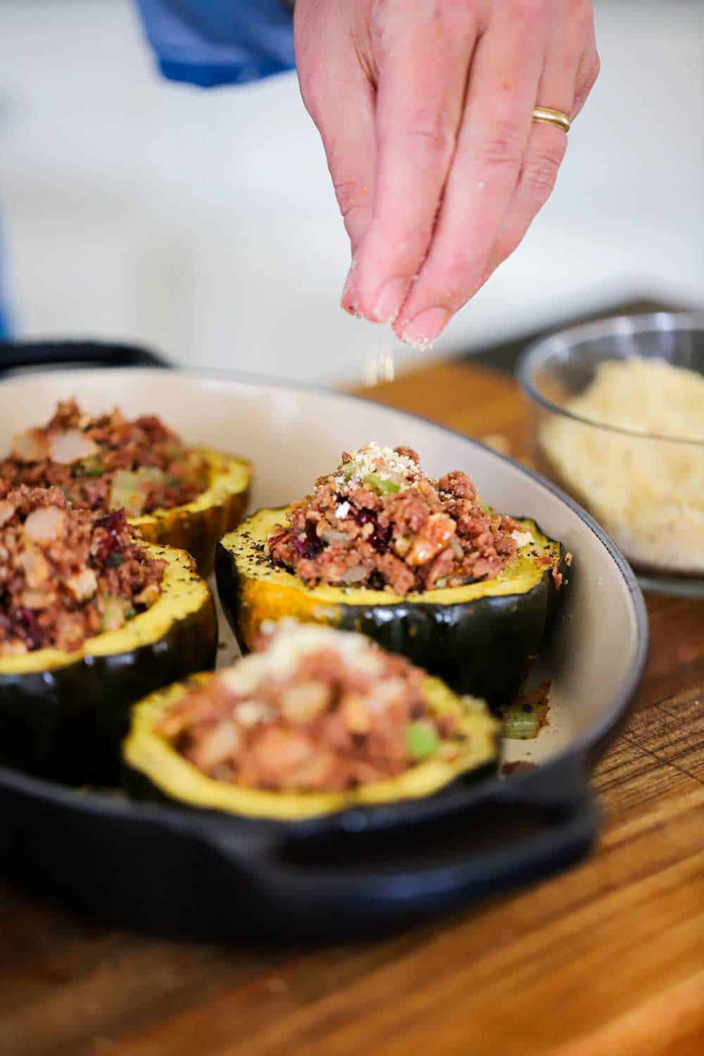 A hand sprinkling bread crumbs onto of stuffed acorn squash in a baking dish.