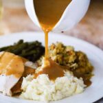A white gravy boat pouring turkey gravy over a plate of mashed potatoes, dressing, turkey, and green beans.