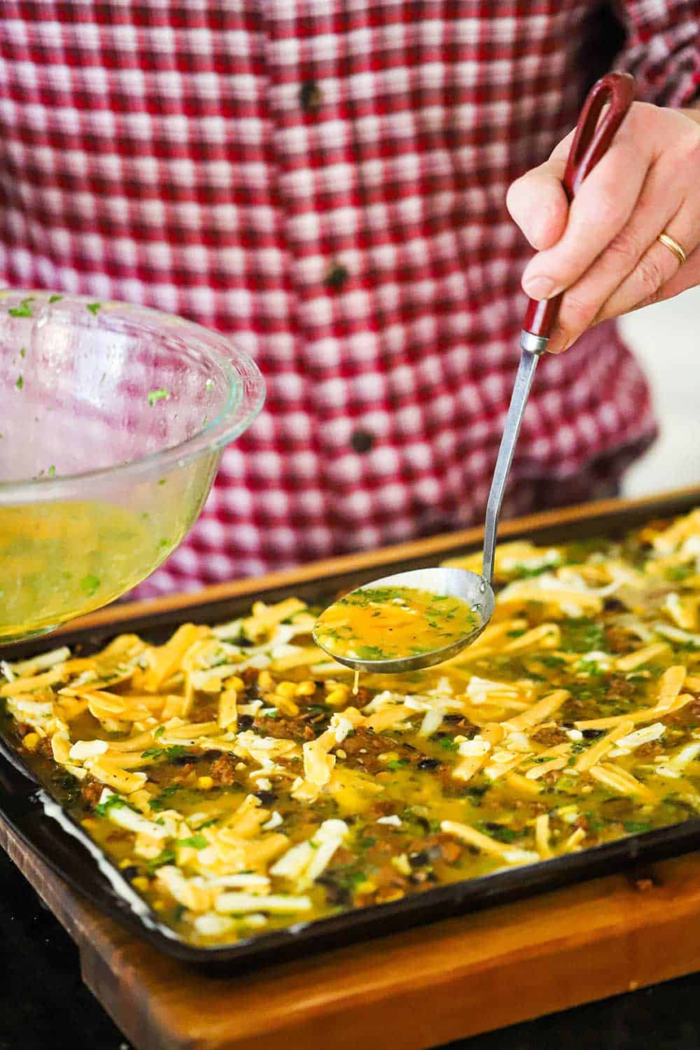 A person using a ladle to spread an egg mixture over the top of a plant-based protein mixture in a rimmed sheet pan.