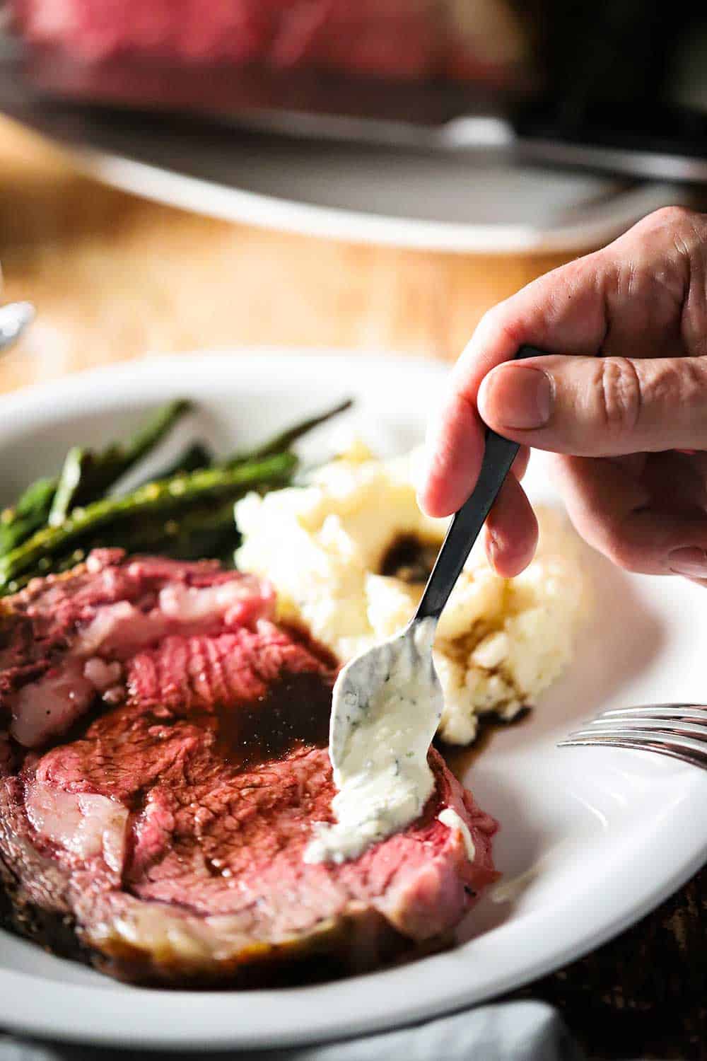 A hand spooning a small dollop of creamy horseradish sauce over a slice of prime rib.