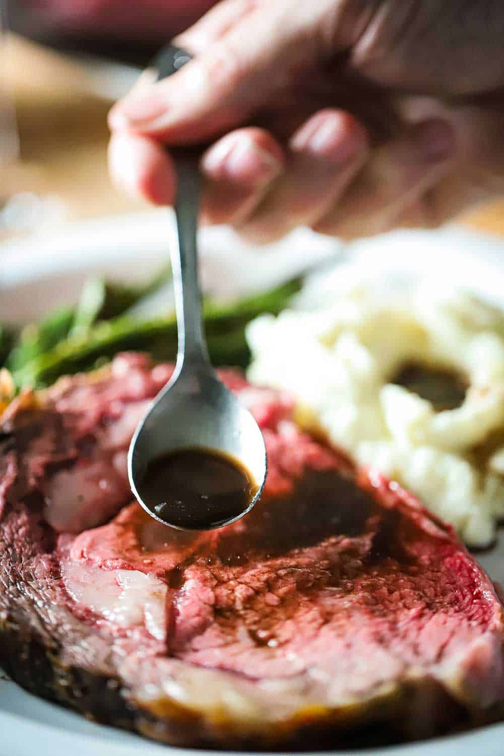 A hand drizzling au jus sauce over a slice of prime rib roast on a plate.