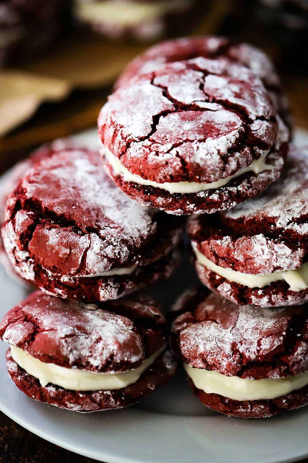 A white plate holding a stack of red velvet sandwich cookies stuffed with cream cheese frosting.