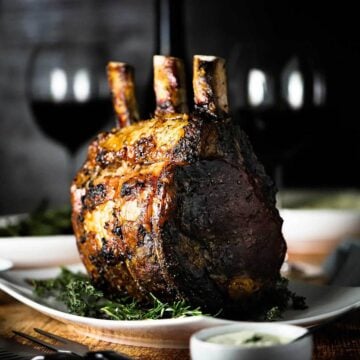 A fully cooked standing prime rib roast sitting upright on a white platter surrounded by fresh herbs.