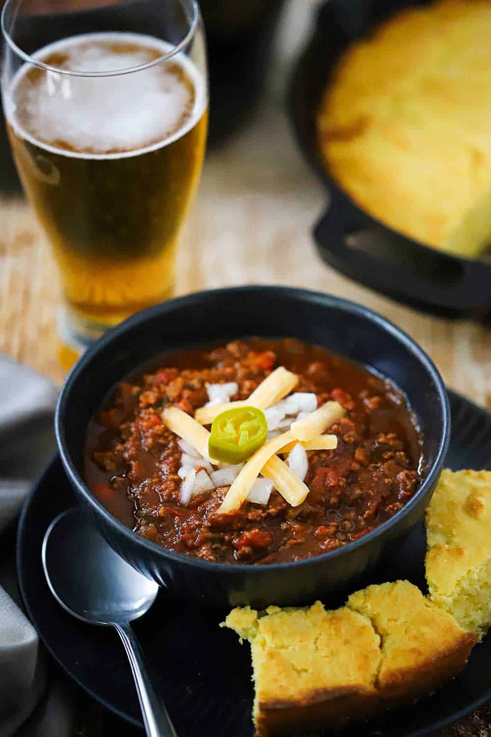 A dark bowl filled with turkey chili topped with shredded cheese and a sliced jalapeno all sitting next to a tall glass of beer.