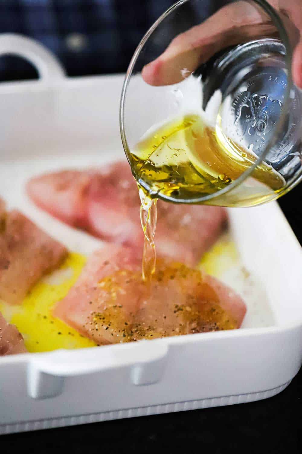 A hand pouring olive oil from a small glass bowl onto a baking dish filled with red snapper fillets.