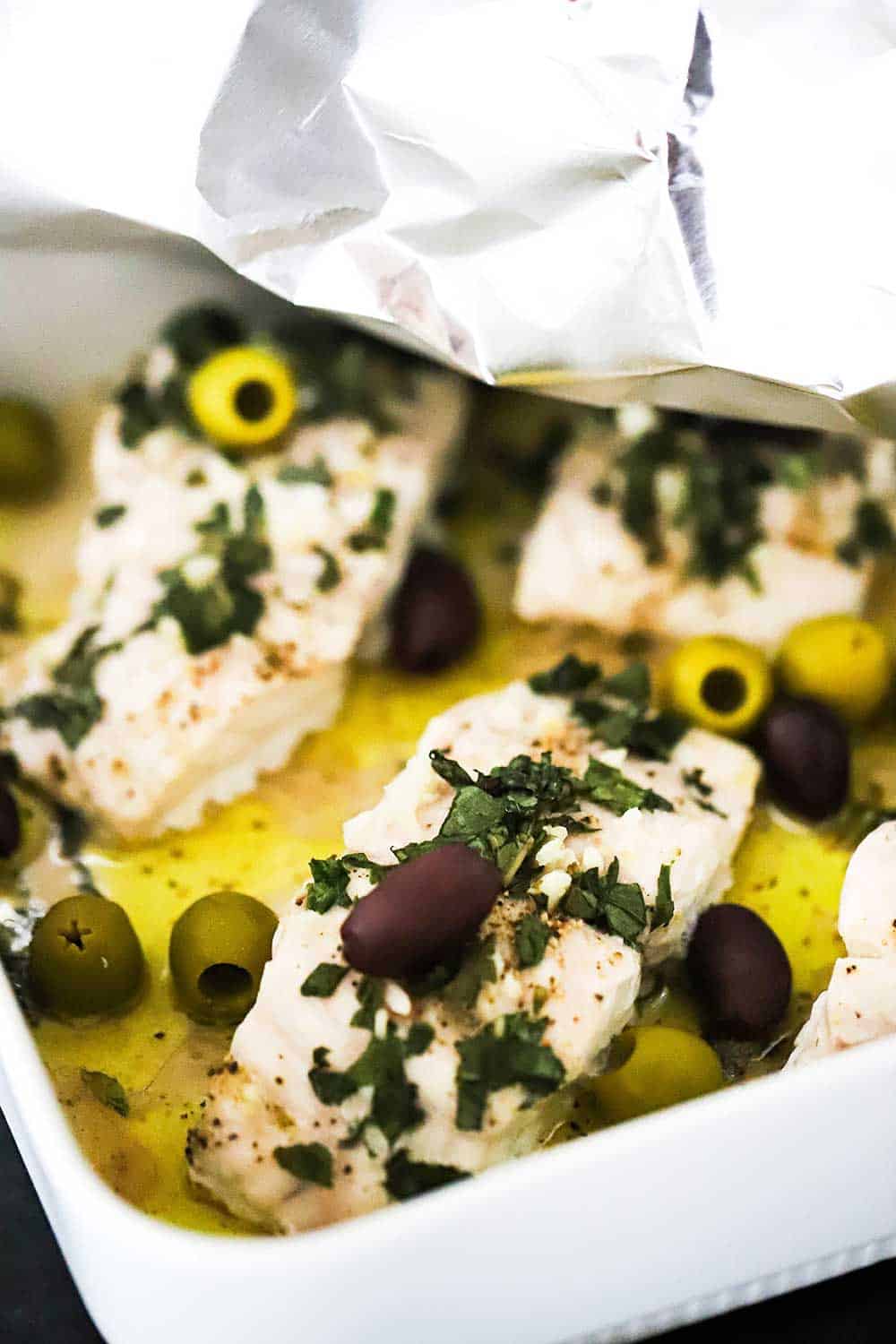 A square white baking dish filled with baked red snapper with a piece of foil being pulled away from the top of the dish.