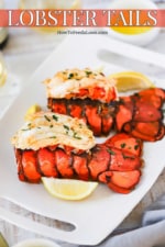 Two fully cooked lobster tails with meat on top of the lobster tail, all sitting on a platter next to lemon wedges and melted butter.