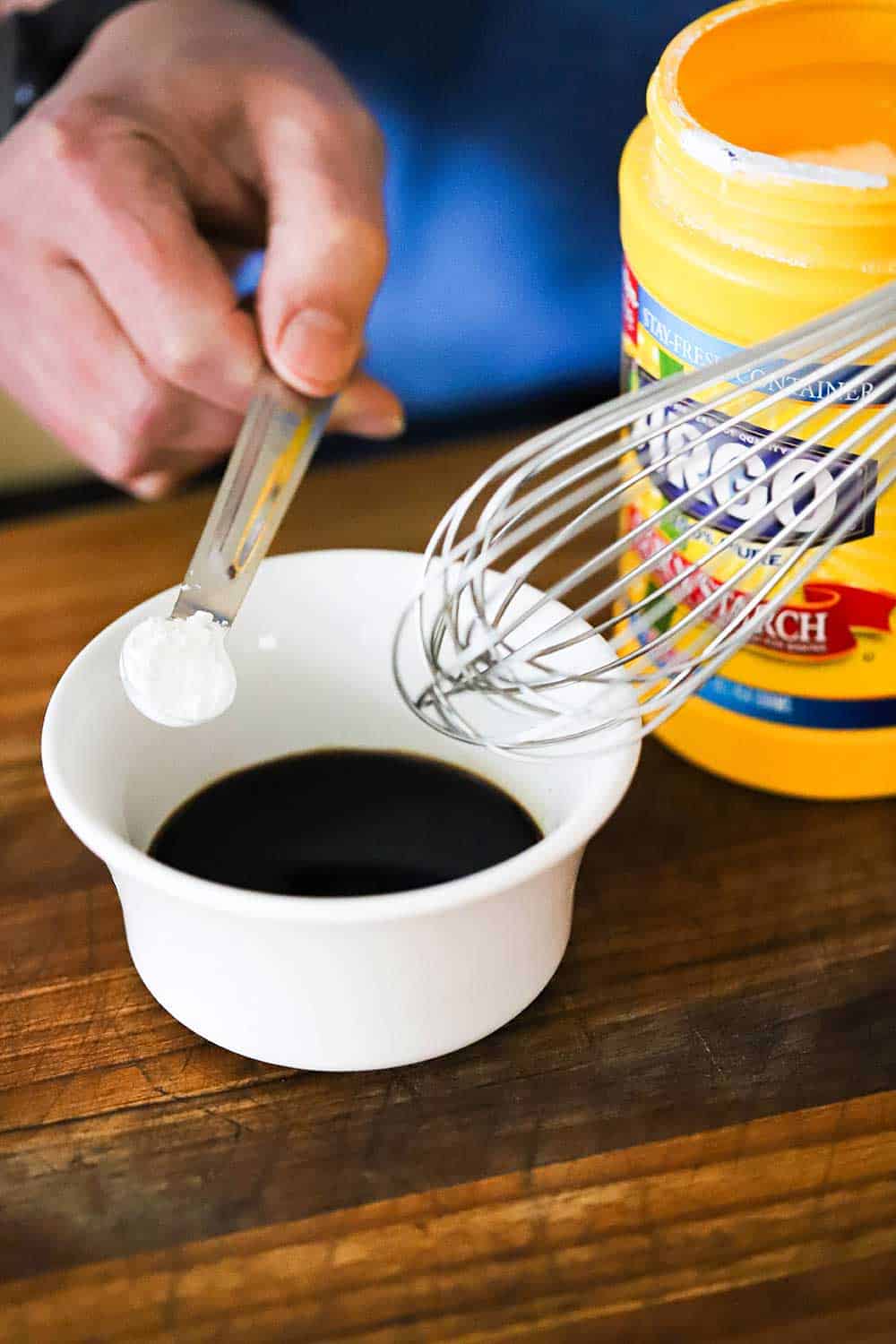 A person holding a quarter-teaspoon filled with cornstarch over a small white bowl filled with soy sauce.