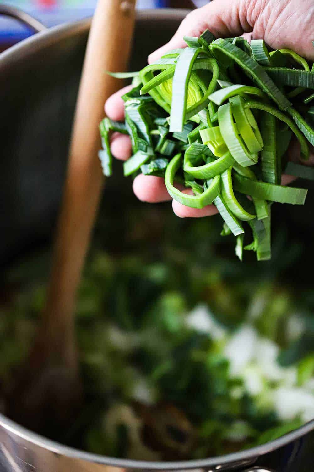 A hand holding sliced leeks over a stock pot filled with chopped onions and celery and wooden spoon in the middle of it.