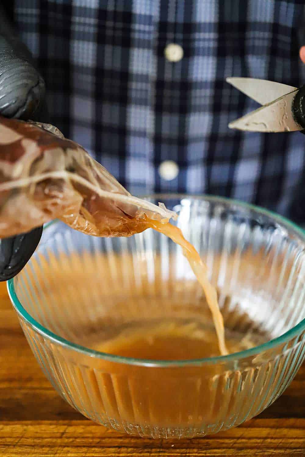 A person holding a bag of cooked short ribs that has been snipped with scissors, allowing the juices to pour into a glass bowl.
