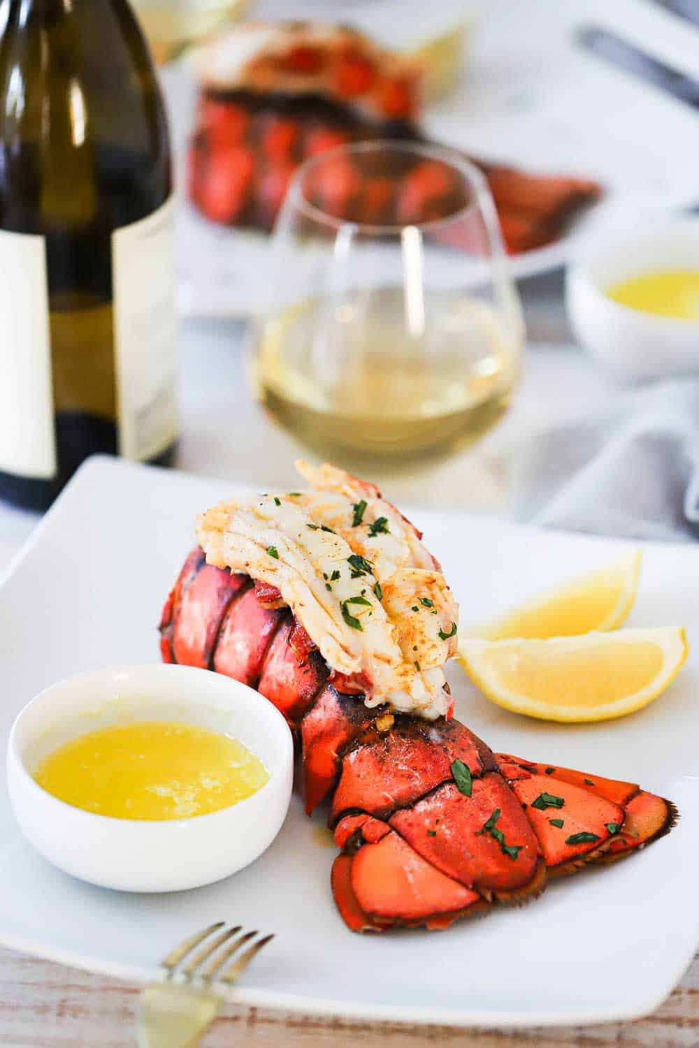 A square white dinner plate filled with a fully cooked lobster tail with a small bowl of drawn butter and lemon wedges next to it, all next to a glass of white wine.