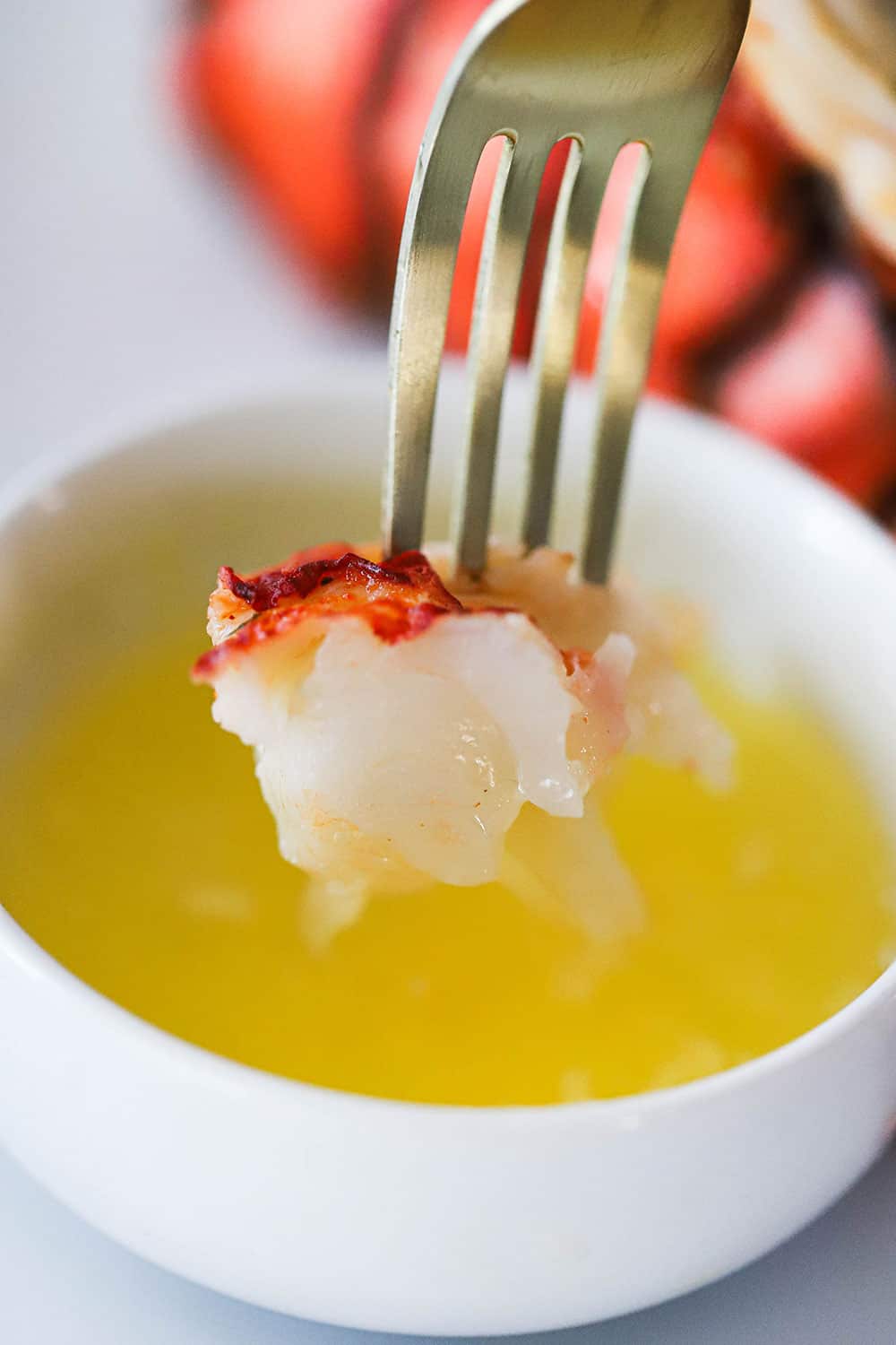 A large piece of cooked lobster meat on the end of a fork that has just been pulled from a bowl of drawn butter.