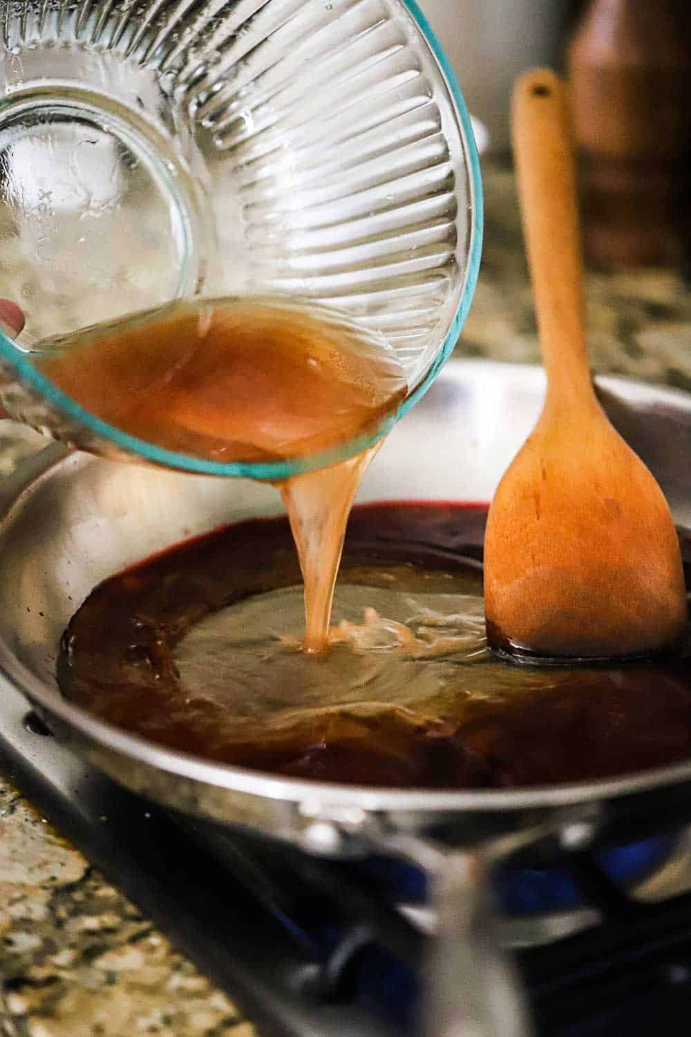 A person pouring beef drippings from a glass bowl into a skillet filled with simmering red wine.