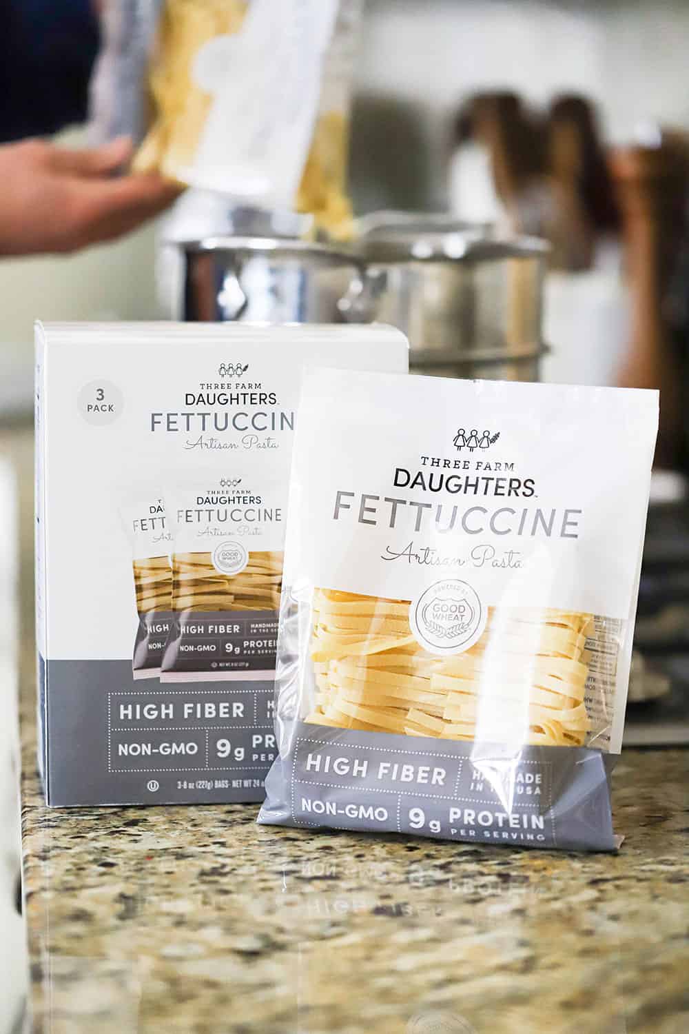 Two packages of dried fettuccine on a counter in front of a pot of boiling water on a stove with a person dropping more of the dried pasta into it.