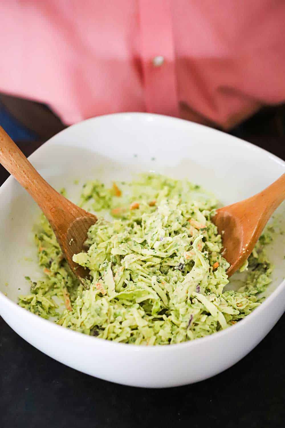 A person in a pink shirt standing behind a white serving bowl filled with avocado-lime slaw with two wooden spoons inserted in it.