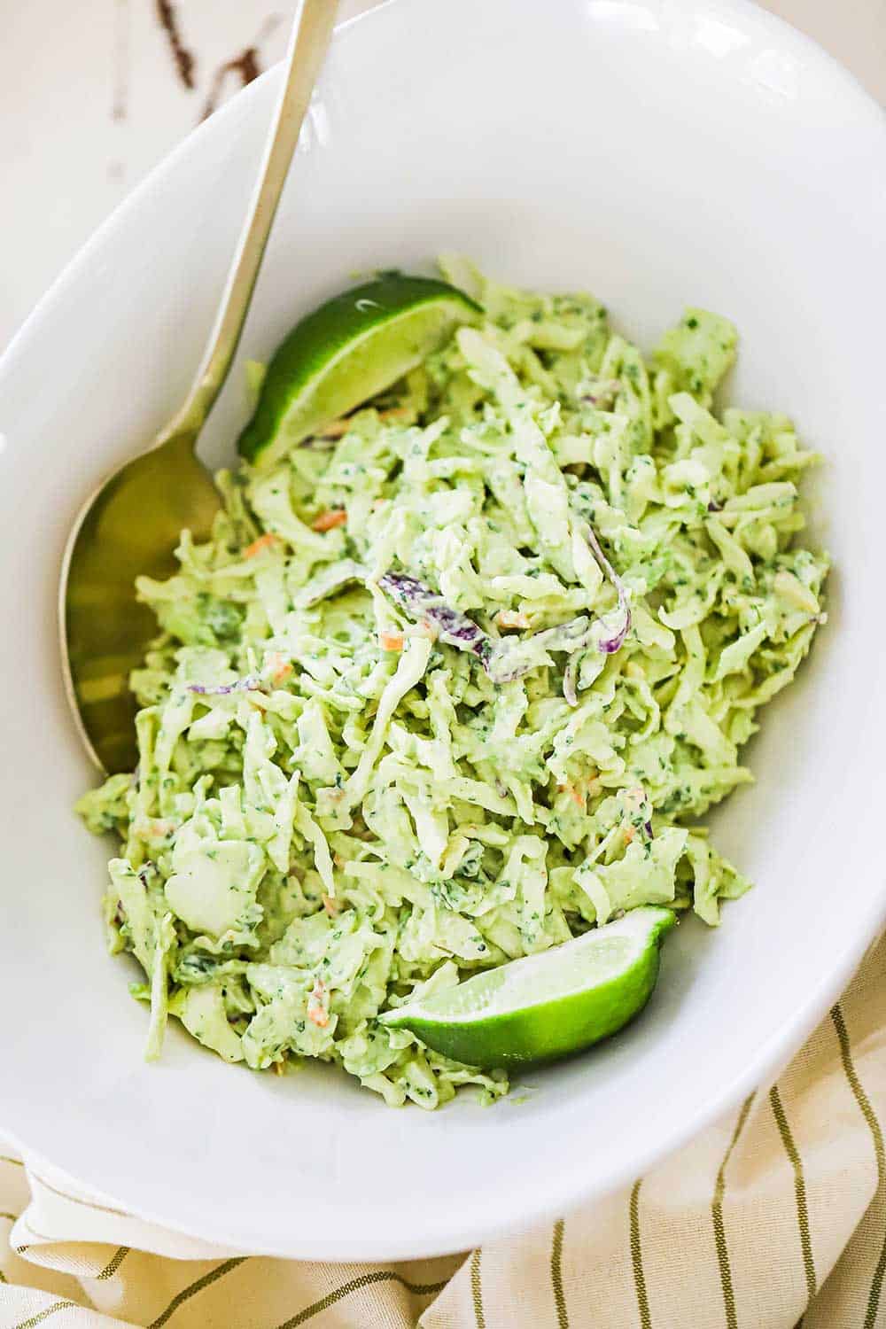 An oval white serving bowl filled with avocado-lime slaw with a gold spoon inserted on the side and a lime wedge on the other side.
