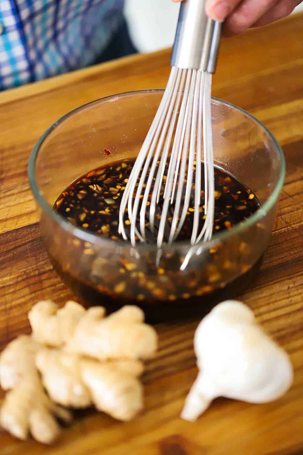 A person using a whisk to mix together a Mongolian beef sauce on a cutting board in front of fresh ginger and a head of garlic.