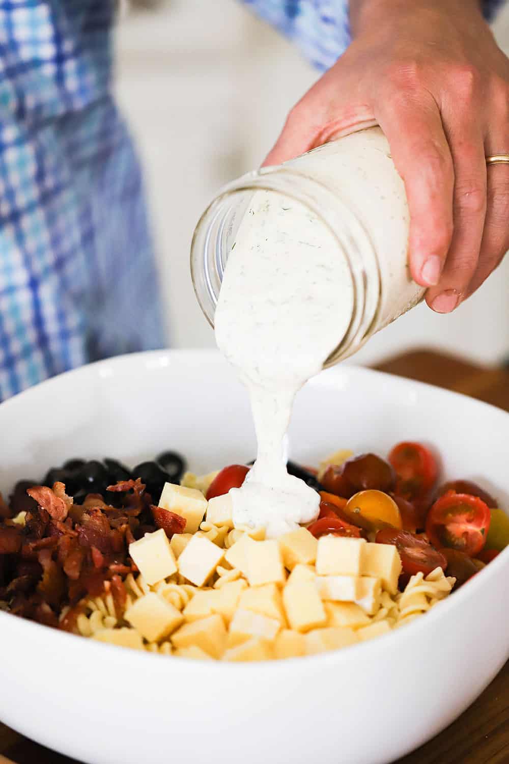 A person pouring homemade Ranch dressing from a glass jar into a bowl filled with cheese, bacon pieces, cherry tomatoes, and sliced black olives, all on top of cooked pasta.
