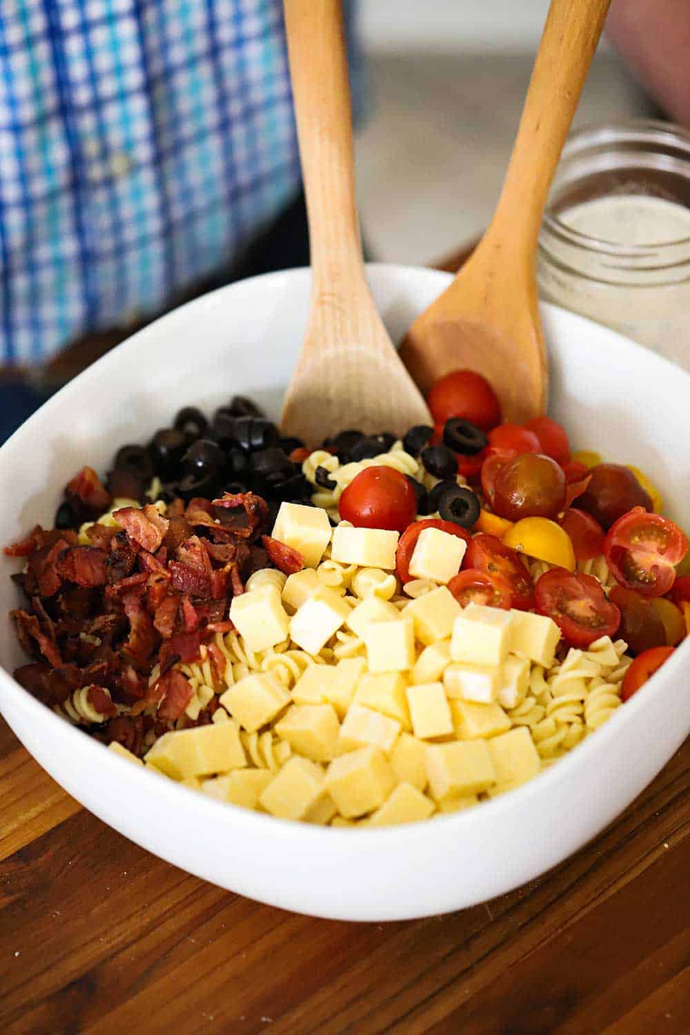 A large white serving bowl filled with pasta topped with cubed cheese, crumbled cooked bacon, sliced black olives, and halved cherry tomatoes.