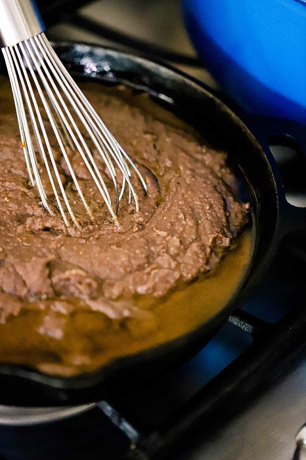 A black cast iron skillet filled with puréed pinto beans that are being whisked with melted lard in the pan.