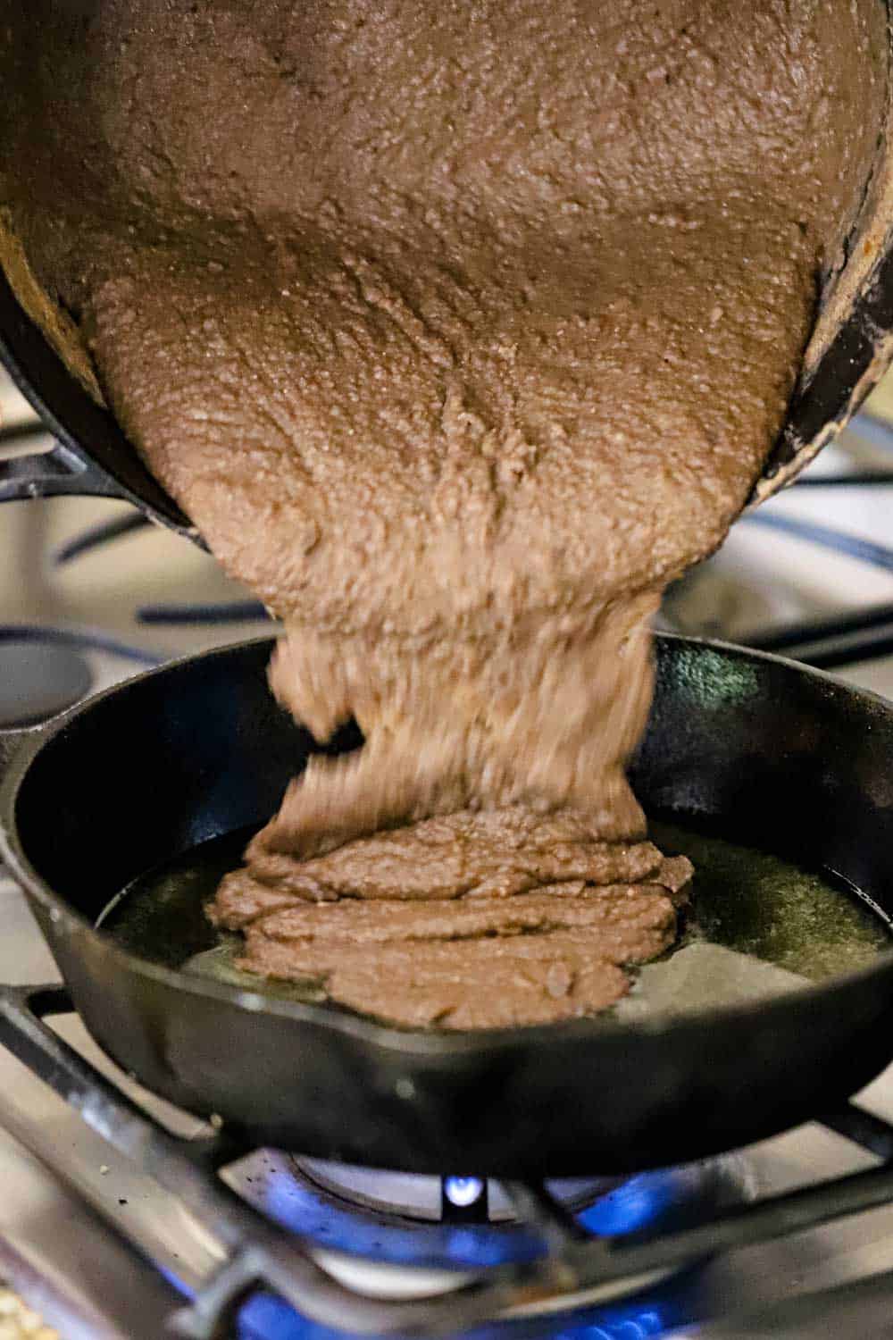 Puréed beans being transferred from a large black pot to a cast iron skillet on a gas stove.