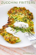 A zucchini fritter that has been split open with a fork and topped with a dollop of sour cream on a white dinner plate.