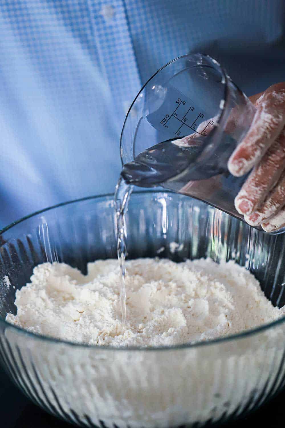 A person pouring water from a clear measuring cup into a glass bowl of sifted flour.