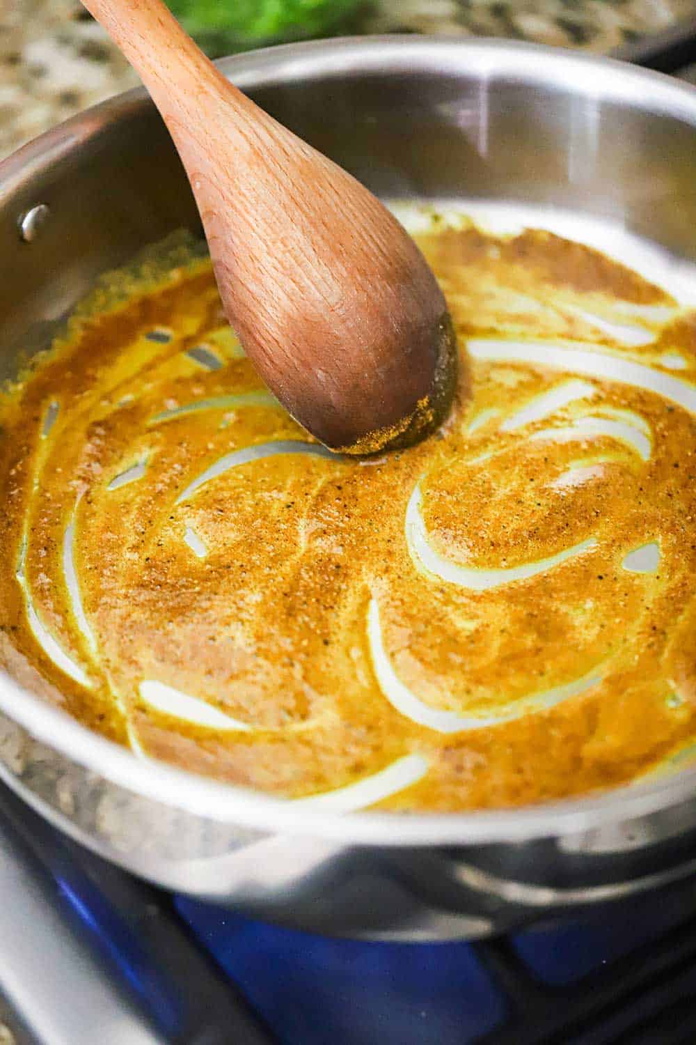 A large stainless steel skillet with melted butter and curry powder in the bottom of the pan with a wooden spoon in the middle of it.