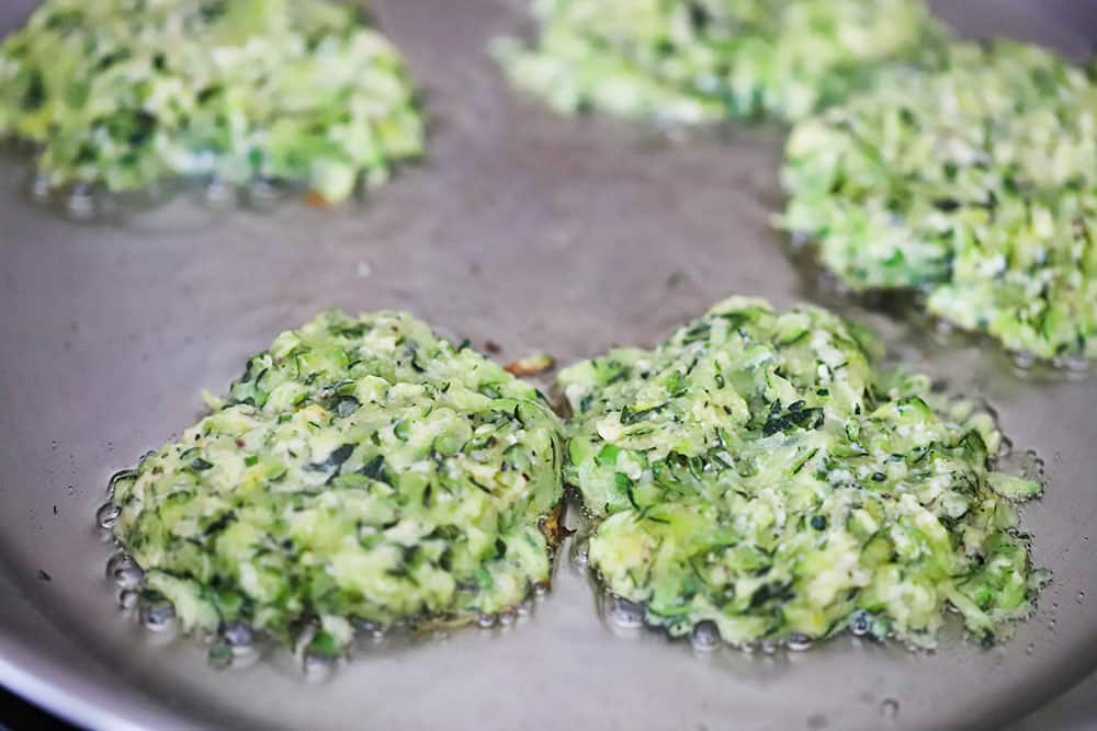 Zucchini fritters being fried in olive oil in a stainless steel skillet.