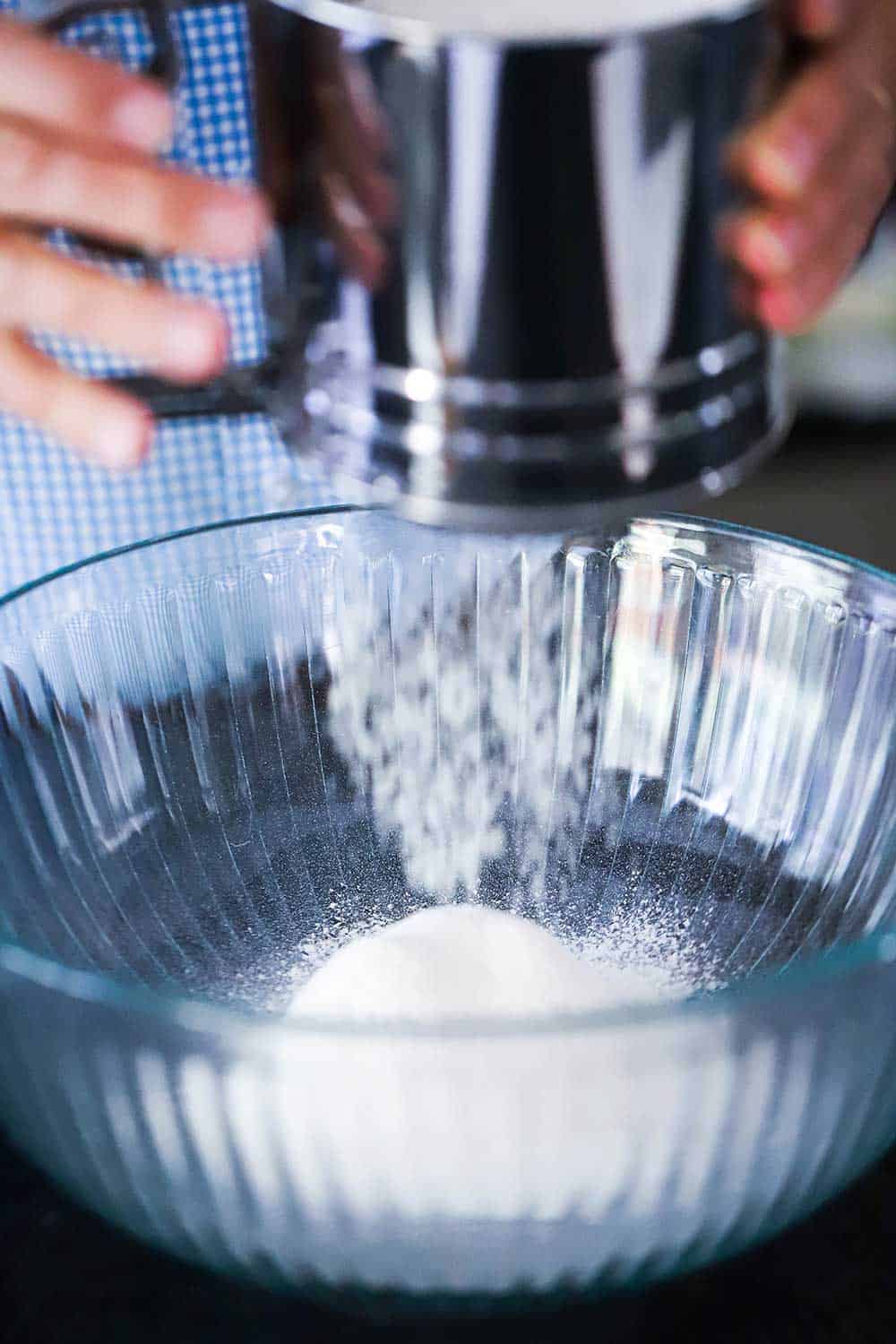 A person holding a metal sifter over a bowl while sifting flour into it.