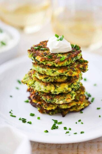 A stack of zucchini fritters on a white dinner plate topped with a dollop of sour cream and snipped chives sprinkled all over.
