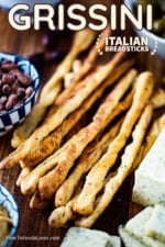 A pile of baked grissini, also known as Italian breadsticks, on a board next to a block of cheese that has been sliced and a bowl of spiced nuts.