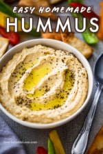 A bowl of homemade hummus with swirls of olive oil and black pepper on top all surrounded by cut bell peppers and pita chips.