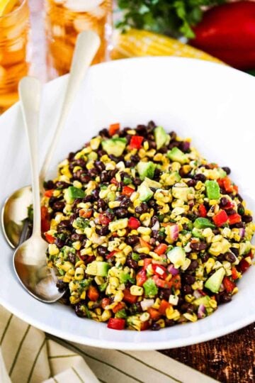A large white serving bowl filled with black bean and corn salad with two gold serving spoons tucked in beside the salad.