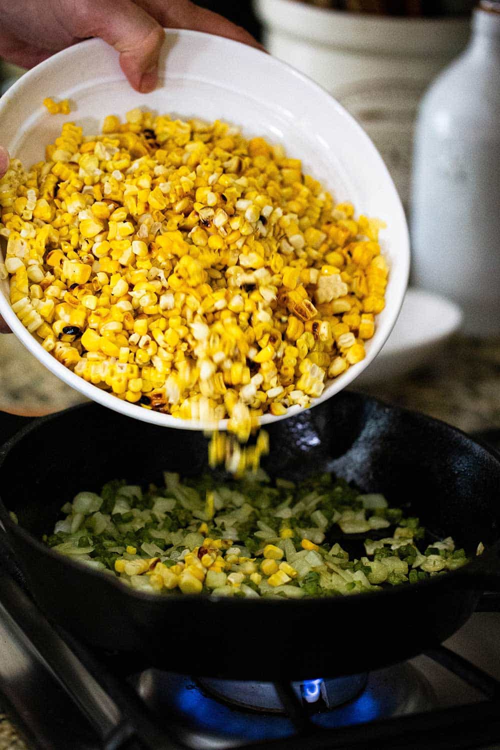 A person dumping grilled kernels of corn into a cast-iron skillet with sautéed onion and peppers in it.