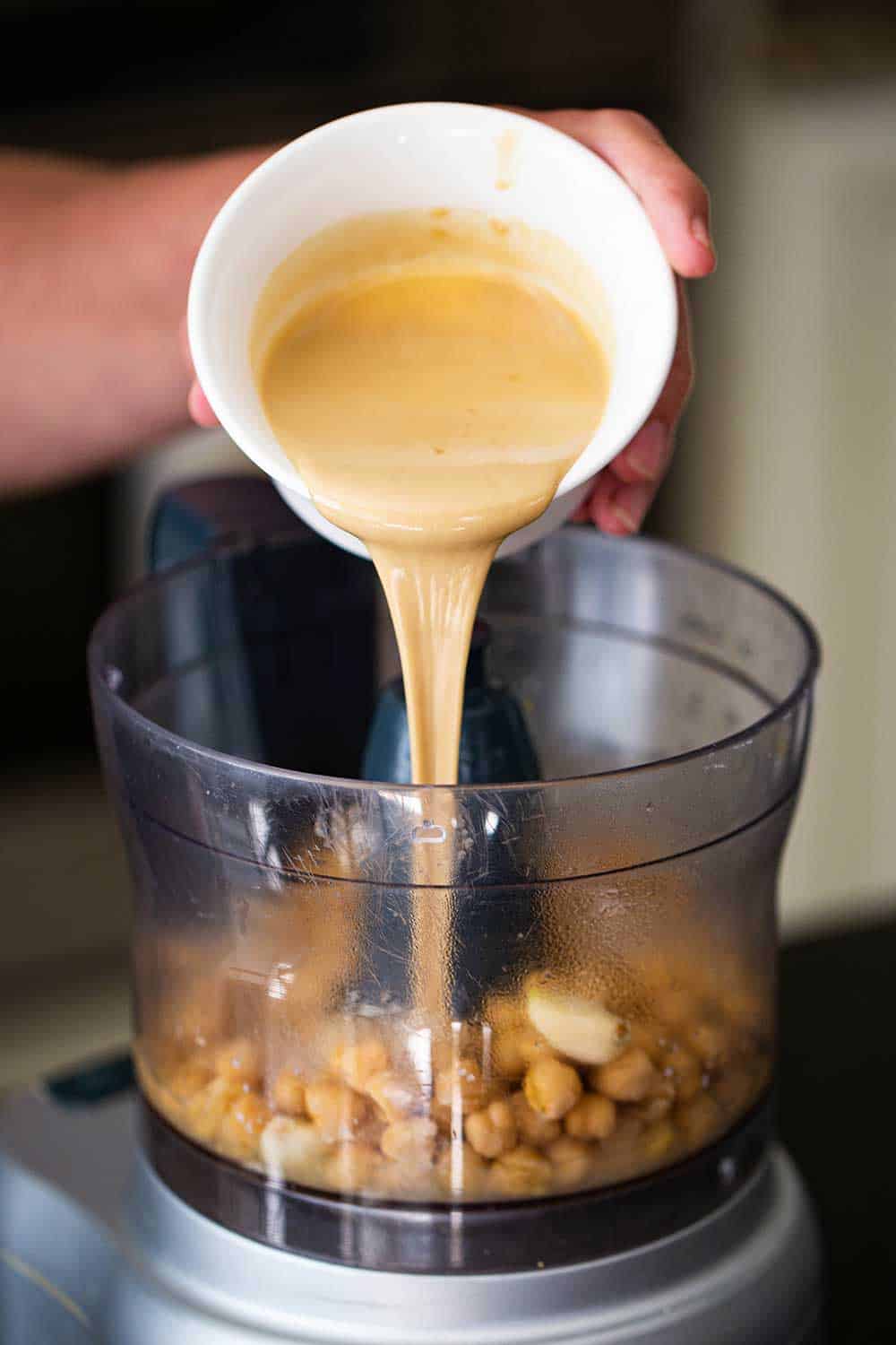 A person pouring tahini sauce from a small white bowl into a bowl filled with cooked chickpeas and garlic.