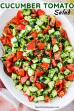 A large white oval serving dish filled with a cucumber tomato salad next to a red checkered napkin.