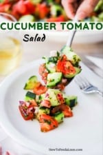 A person holding a large serving spoon filled with cucumber tomato salad over a white dinner plate with a helping of the salad on it.