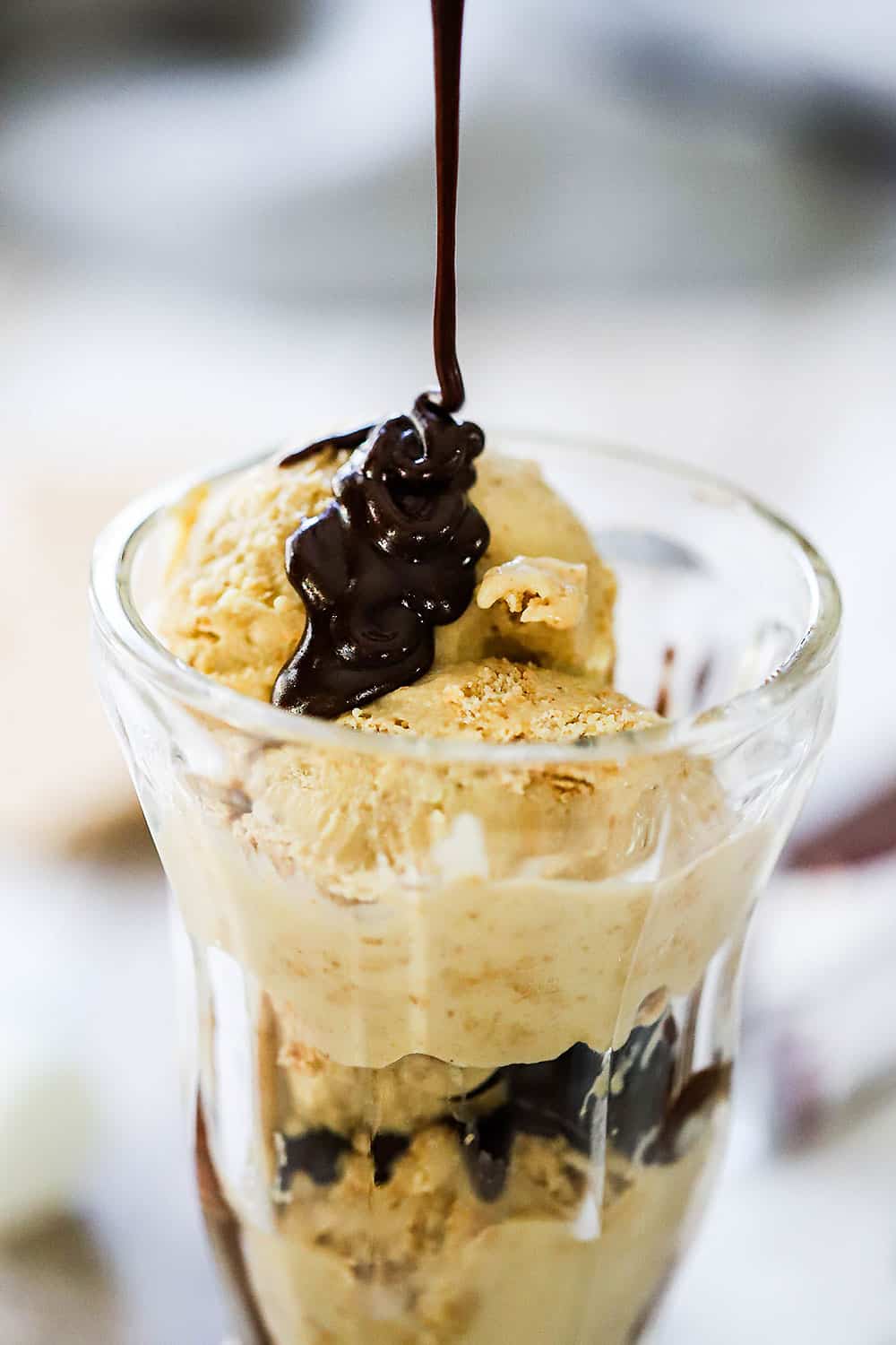 A stream of chocolate sauce being poured onto an ice cream glass holder filled with s'mores ice cream.