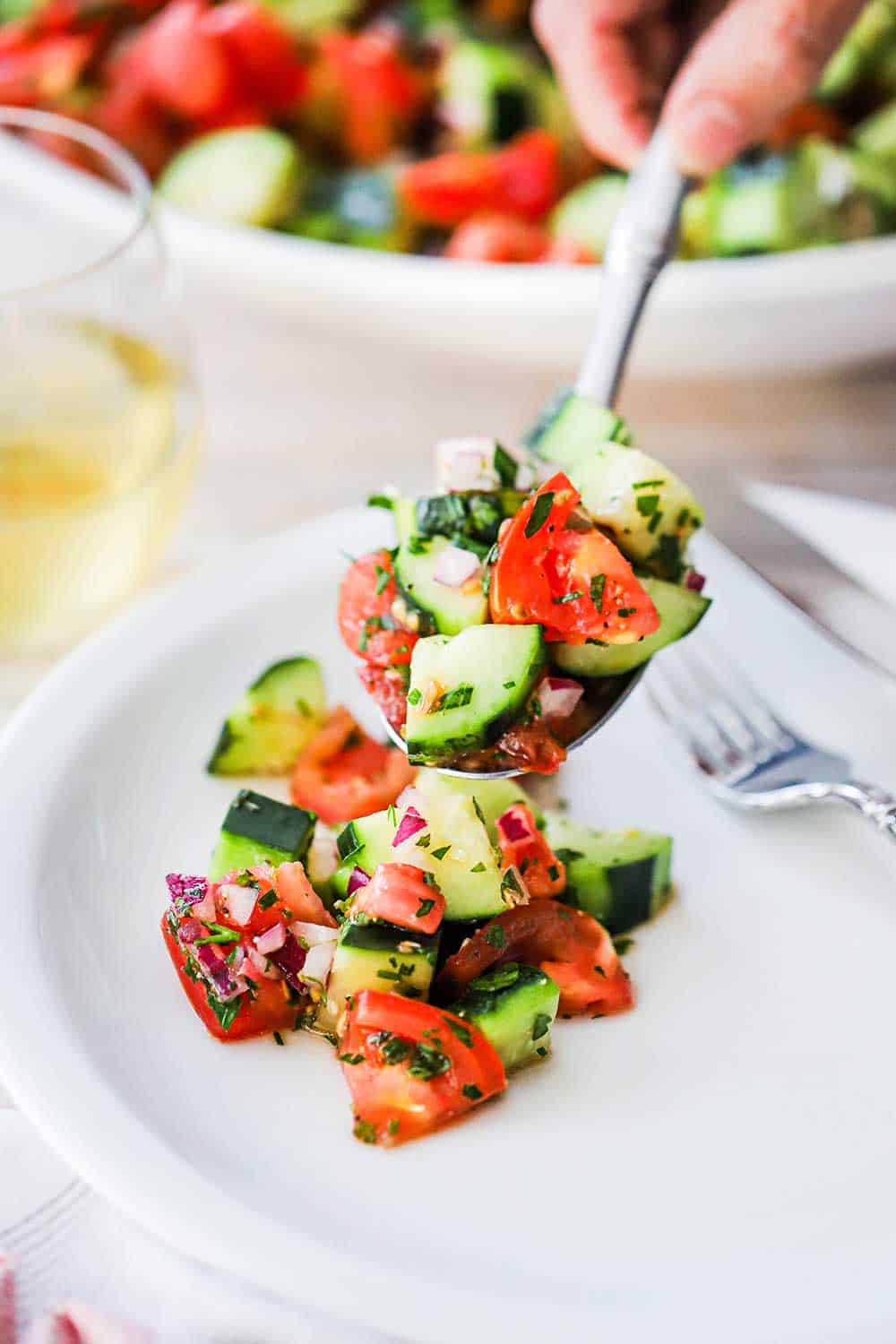 A person holding a large serving spoon filled with cucumber tomato salad over a white dinner plate with a helping of the salad on it.