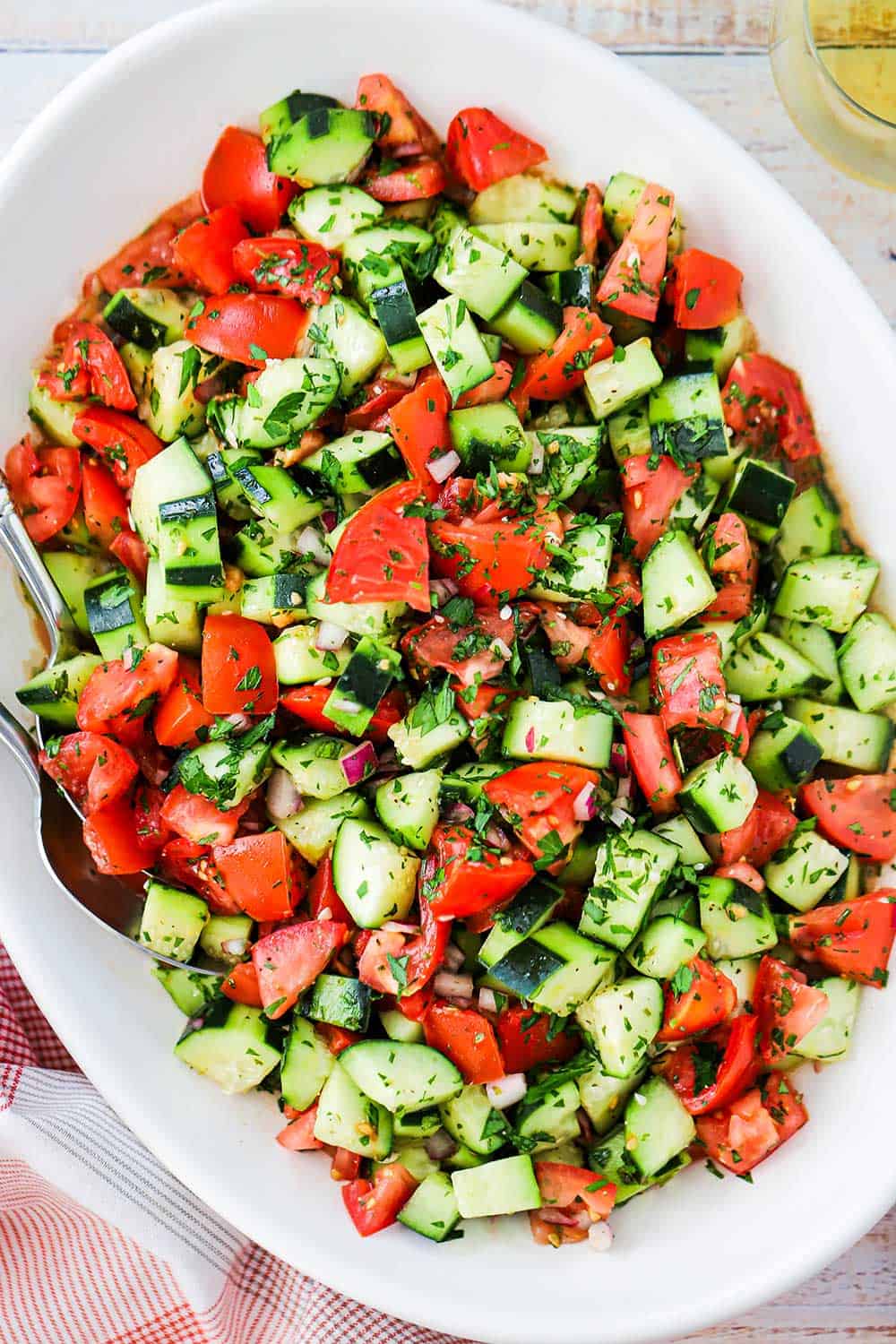 A large white oval serving dish filled with a cucumber tomato salad next to a red checkered napkin.