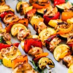 A row of 5 chicken kabobs on a white oval platter garnish with fresh rosemary sprigs and a bowl of white sauce.