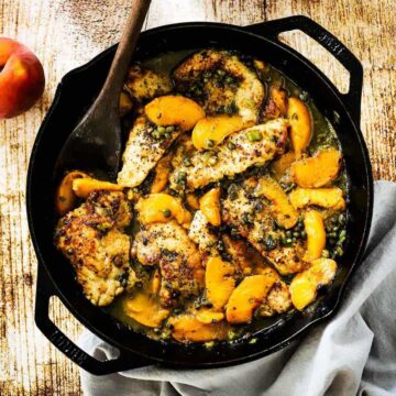 A large black cast-iron skillet filled with pan-fried pork cutlets and peaches in a savory sauce all next to several whole peaches.