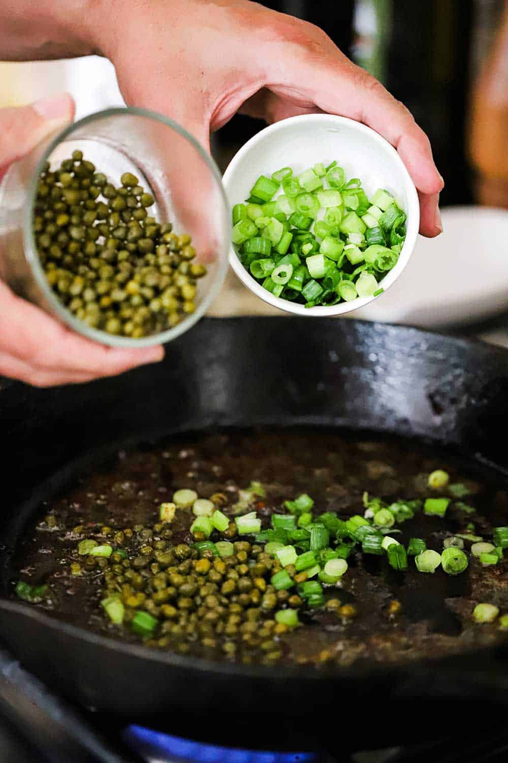 A person dumping capers with one hand from a small bowl, and the other hand dumping chopped scallions, both into a cast-iron skillet. over a cast-iron skillet