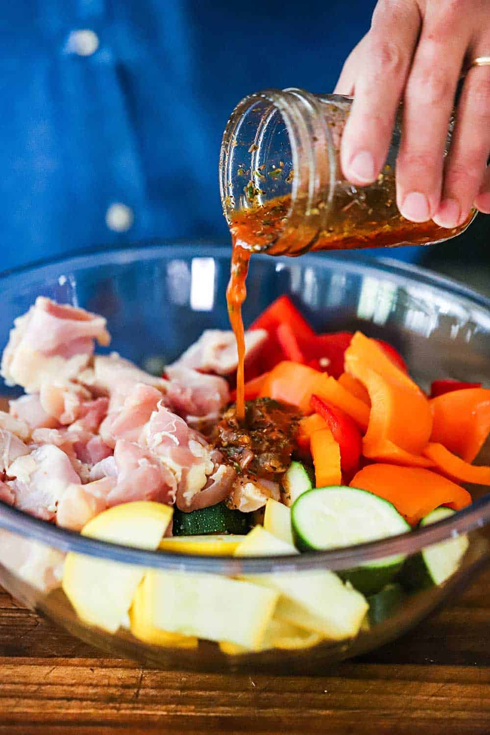 A person pouring a red wine vinegar marinade over cut up chicken and colorful vegetables in a glass bowl.