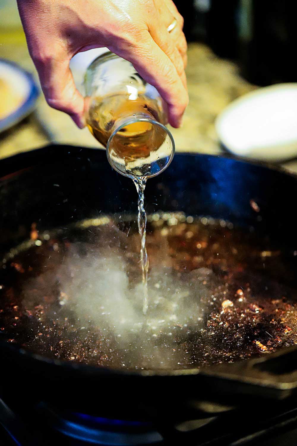 A person pouring sherry from a small carafe into a cast-iron skillet with pieces of breading stuck to the surface of the pan.